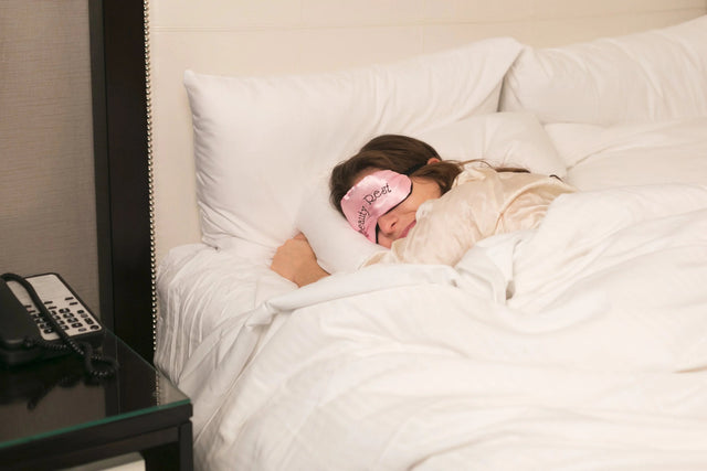 Woman sleeping in a cozy bed with white bedding, wearing a pink sleep mask, enjoying restful sleep